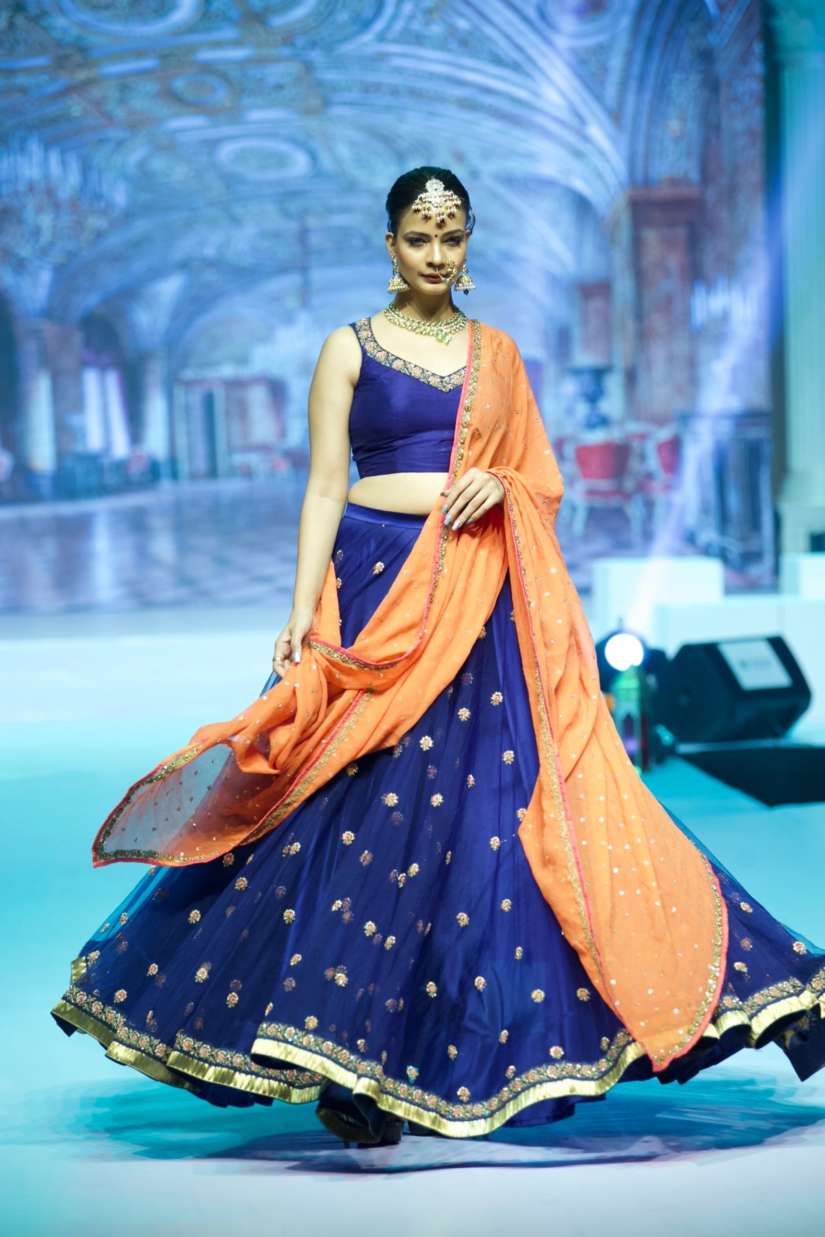 Model in a navy lehenga with an orange dupatta, mid-twirl